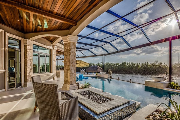 elevated and luxurious outdoor patio with vaulted wooden ceilings overlooking a glass enclosure over an infinity pool on a lake